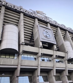 Tall structural facade of a large stadium with visible columns and a silver-gray exterior. The signage reads 'Estadio Santiago Bernabéu' and there is a prominent logo of Real Madrid Club de Fútbol displayed.
