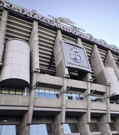 Tall structural facade of a large stadium with visible columns and a silver-gray exterior. The signage reads 'Estadio Santiago Bernabéu' and there is a prominent logo of Real Madrid Club de Fútbol displayed.