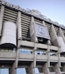 Tall structural facade of a large stadium with visible columns and a silver-gray exterior. The signage reads 'Estadio Santiago Bernab&eacute;u' and there is a prominent logo of Real Madrid Club de F&uacute;tbol displayed.