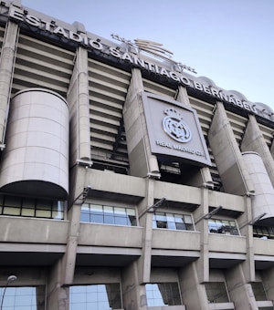 Tall structural facade of a large stadium with visible columns and a silver-gray exterior. The signage reads 'Estadio Santiago Bernab&eacute;u' and there is a prominent logo of Real Madrid Club de F&uacute;tbol displayed.
