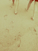 Smiling travelers walking barefoot along a sandy shore with footprints behind them.