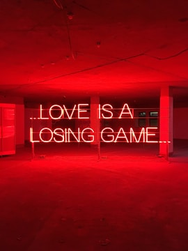 Red neon lights form the words 'LOVE IS A LOSING GAME' in an empty room. The room has a concrete floor and a ceiling with visible beams and light fixtures.