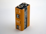A close-up view of a rectangular 9V battery with an orange and black design. The battery is labeled 'Industrial by Duracell' and 'Professional Alkaline Battery.' Various icons and text, including production details and recycling information, are printed on the surface.