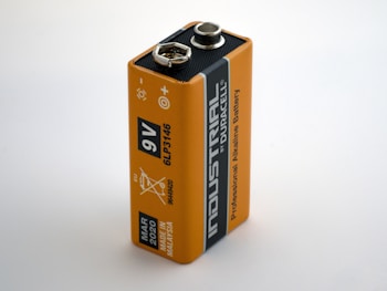A close-up view of a rectangular 9V battery with an orange and black design. The battery is labeled 'Industrial by Duracell' and 'Professional Alkaline Battery.' Various icons and text, including production details and recycling information, are printed on the surface.
