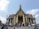 A large, ornately decorated temple with golden embellishments and intricate architecture stands against a clear blue sky. Many people are gathered on the steps leading up to the temple entrance, some holding umbrellas and taking photos. The roof features traditional Thai design elements with sharp, tiered edges.