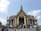 A large, ornately decorated temple with golden embellishments and intricate architecture stands against a clear blue sky. Many people are gathered on the steps leading up to the temple entrance, some holding umbrellas and taking photos. The roof features traditional Thai design elements with sharp, tiered edges.