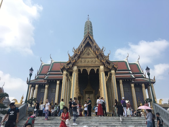 A large, ornately decorated temple with golden embellishments and intricate architecture stands against a clear blue sky. Many people are gathered on the steps leading up to the temple entrance, some holding umbrellas and taking photos. The roof features traditional Thai design elements with sharp, tiered edges.