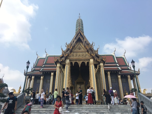 A large, ornately decorated temple with golden embellishments and intricate architecture stands against a clear blue sky. Many people are gathered on the steps leading up to the temple entrance, some holding umbrellas and taking photos. The roof features traditional Thai design elements with sharp, tiered edges.