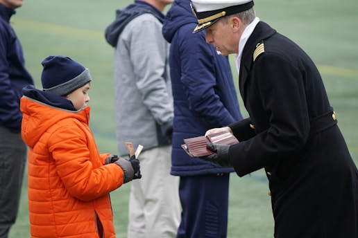 A person in a naval uniform is interacting with a child wearing a bright orange jacket and a blue beanie. They are exchanging items, possibly cards or papers. Other people stand in the background on a field with green turf.