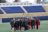 A group of female lacrosse players huddle together on a field, raising their lacrosse sticks in the air. They are wearing matching uniforms in red and black, with several in winter jackets. The field is situated in a stadium with empty blue seats and a lacrosse goal visible in the background.