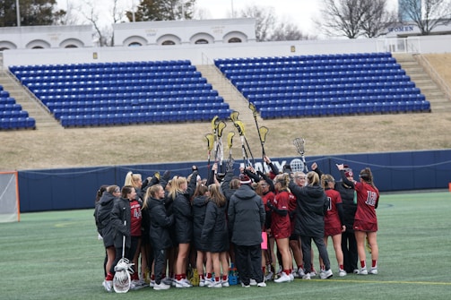 A group of female lacrosse players huddle together on a field, raising their lacrosse sticks in the air. They are wearing matching uniforms in red and black, with several in winter jackets. The field is situated in a stadium with empty blue seats and a lacrosse goal visible in the background.