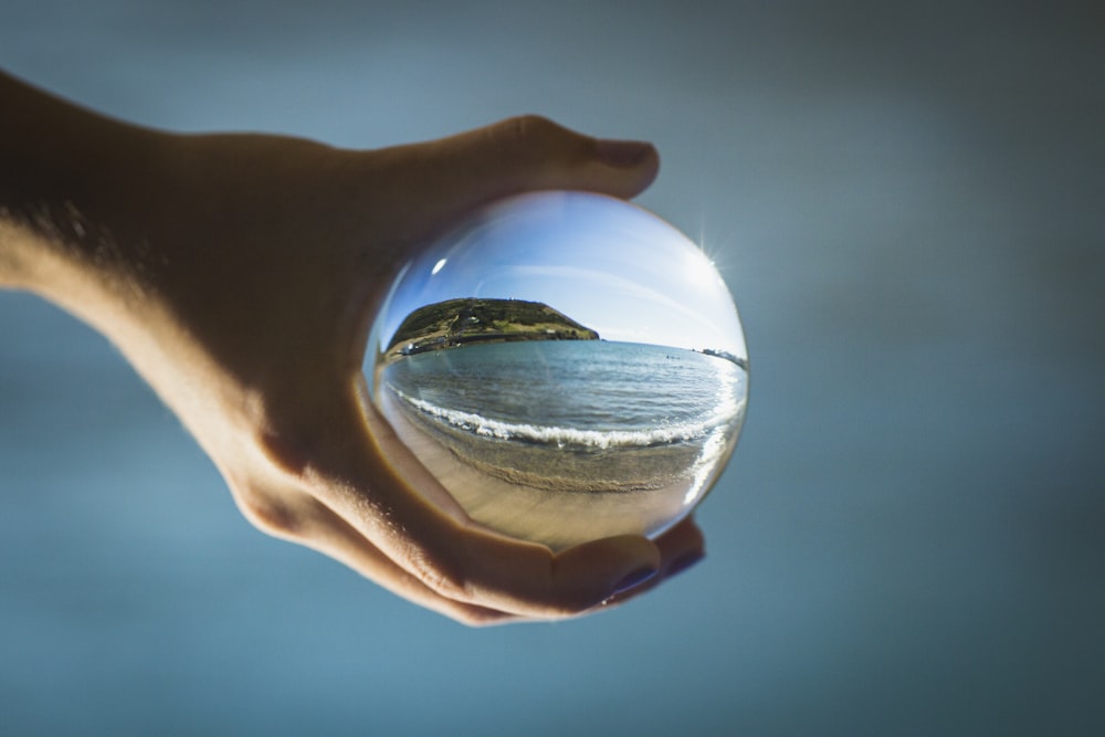 pessoa segurando bola de vidro refletindo o mar durante o dia