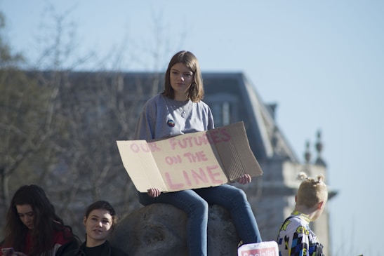A group of young people is gathered outdoors with a large building in the background. One person is sitting on a stone holding a cardboard sign that reads 'OUR FUTURE IS ON THE LINE.' The atmosphere appears calm and sunny, with some trees in the blurred background.