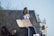 A group of young people is gathered outdoors with a large building in the background. One person is sitting on a stone holding a cardboard sign that reads 'OUR FUTURE IS ON THE LINE.' The atmosphere appears calm and sunny, with some trees in the blurred background.
