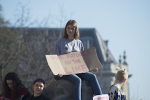 A group of young people is gathered outdoors with a large building in the background. One person is sitting on a stone holding a cardboard sign that reads 'OUR FUTURE IS ON THE LINE.' The atmosphere appears calm and sunny, with some trees in the blurred background.