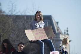 A group of young people is gathered outdoors with a large building in the background. One person is sitting on a stone holding a cardboard sign that reads 'OUR FUTURE IS ON THE LINE.' The atmosphere appears calm and sunny, with some trees in the blurred background.