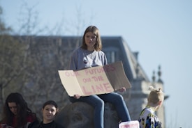 A group of young people is gathered outdoors with a large building in the background. One person is sitting on a stone holding a cardboard sign that reads 'OUR FUTURE IS ON THE LINE.' The atmosphere appears calm and sunny, with some trees in the blurred background.