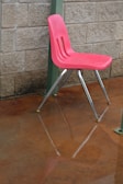 A red plastic chair with metal legs is positioned on a wet concrete floor. The chair is slightly elevated from the ground, and its reflection is visible on the shiny surface below. The background features a textured beige brick wall with a green metal pillar on the side.