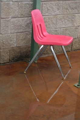 A red plastic chair with metal legs is positioned on a wet concrete floor. The chair is slightly elevated from the ground, and its reflection is visible on the shiny surface below. The background features a textured beige brick wall with a green metal pillar on the side.