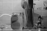A kitchen countertop displays a collection of utensils in a glass container, including wooden spoons and spatulas. Nearby is a large glass jar filled with water, possibly containing cucumber slices, and a smaller bowl with spoons and corks. The background shows tiles with coffee-related decor.
