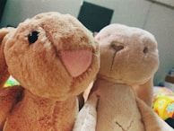 Two plush toy animals are positioned close together. One is a light brown rabbit with a prominent pink nose and black eyes, while the other is a beige rabbit with a subtle smile and darker stitching for facial features. The background is softly blurred, hinting at colorful patterns.