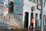 A street scene with a person in an orange reflective vest and red cap cleaning the pavement. The building features multiple doors and windows with ornate metalwork. The walls are adorned with colorful graffiti, and there are visible signs of wear and tear on the structure.