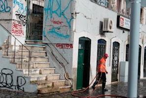 A street scene with a person in an orange reflective vest and red cap cleaning the pavement. The building features multiple doors and windows with ornate metalwork. The walls are adorned with colorful graffiti, and there are visible signs of wear and tear on the structure.