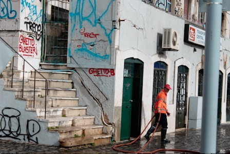 A street scene with a person in an orange reflective vest and red cap cleaning the pavement. The building features multiple doors and windows with ornate metalwork. The walls are adorned with colorful graffiti, and there are visible signs of wear and tear on the structure.