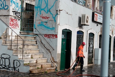 A street scene with a person in an orange reflective vest and red cap cleaning the pavement. The building features multiple doors and windows with ornate metalwork. The walls are adorned with colorful graffiti, and there are visible signs of wear and tear on the structure.