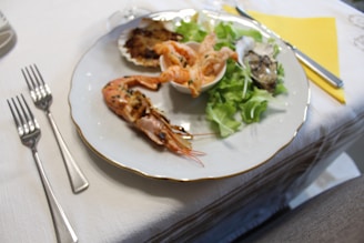 A white plate is elegantly arranged with seafood including prawns and oysters, accompanied by a small portion of salad greens. The setting includes shiny cutlery on a white tablecloth and a yellow napkin.
