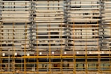 A building facade is under construction or renovation, featuring a complex pattern of horizontal and vertical lines. The exterior structure includes scaffolding in the foreground, characterized by its grid-like arrangement of yellow poles. The facade displays a series of horizontal slats, windows, and air conditioning units.