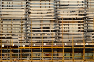 A building facade is under construction or renovation, featuring a complex pattern of horizontal and vertical lines. The exterior structure includes scaffolding in the foreground, characterized by its grid-like arrangement of yellow poles. The facade displays a series of horizontal slats, windows, and air conditioning units.