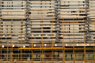 A building facade is under construction or renovation, featuring a complex pattern of horizontal and vertical lines. The exterior structure includes scaffolding in the foreground, characterized by its grid-like arrangement of yellow poles. The facade displays a series of horizontal slats, windows, and air conditioning units.