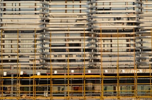 A building facade is under construction or renovation, featuring a complex pattern of horizontal and vertical lines. The exterior structure includes scaffolding in the foreground, characterized by its grid-like arrangement of yellow poles. The facade displays a series of horizontal slats, windows, and air conditioning units.