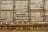 A building facade is under construction or renovation, featuring a complex pattern of horizontal and vertical lines. The exterior structure includes scaffolding in the foreground, characterized by its grid-like arrangement of yellow poles. The facade displays a series of horizontal slats, windows, and air conditioning units.