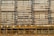 A building facade is under construction or renovation, featuring a complex pattern of horizontal and vertical lines. The exterior structure includes scaffolding in the foreground, characterized by its grid-like arrangement of yellow poles. The facade displays a series of horizontal slats, windows, and air conditioning units.