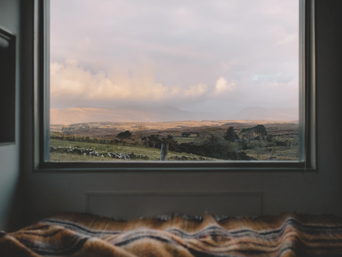 A warm striped blanket draped on a bed in the foreground, with a big window looking out onto rolling hills and a soft cloudy sky — the seasons change outside, the comforter stays perfect inside