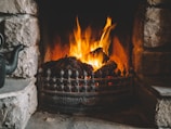 A warm and inviting fireplace with a bright, roaring fire, surrounded by a rustic stone setting. A black teapot is placed on the left side of the hearth, adding a cozy touch to the scene.