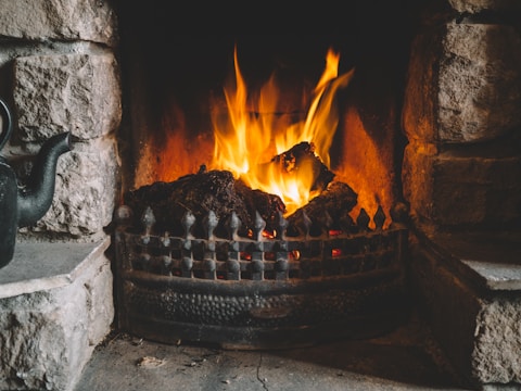 A warm and inviting fireplace with a bright, roaring fire, surrounded by a rustic stone setting. A black teapot is placed on the left side of the hearth, adding a cozy touch to the scene.