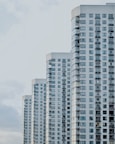 A series of modern high-rise apartment buildings with numerous windows and balconies, positioned side by side in a slightly staggered formation. The buildings appear to be well-maintained and have a uniform architectural style with a clean, minimalistic design.