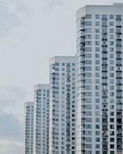 A series of modern high-rise apartment buildings with numerous windows and balconies, positioned side by side in a slightly staggered formation. The buildings appear to be well-maintained and have a uniform architectural style with a clean, minimalistic design.
