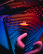 A close-up of a smartphone featuring an apple logo, reflecting vibrant neon lights. The image includes a pair of earbuds and part of a keyboard, all bathed in a dramatic blend of red, blue, and purple hues.
