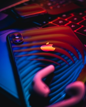 A close-up of a smartphone featuring an apple logo, reflecting vibrant neon lights. The image includes a pair of earbuds and part of a keyboard, all bathed in a dramatic blend of red, blue, and purple hues.