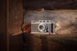 A vintage camera with a textured black and silver body is placed on a wooden surface, surrounded by a few scattered red berries and a dried leaf. The background consists of weathered wooden planks, adding a rustic feel to the scene. Soft, natural light highlights the details of the camera and the surrounding objects.