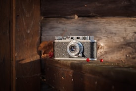 A vintage camera with a textured black and silver body is placed on a wooden surface, surrounded by a few scattered red berries and a dried leaf. The background consists of weathered wooden planks, adding a rustic feel to the scene. Soft, natural light highlights the details of the camera and the surrounding objects.