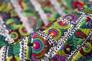 An intricate textile with vibrant embroidery featuring circular and spiral patterns in various colors. The fabric displays a complex design with layers of green, red, purple, and blue threads against a textured background.