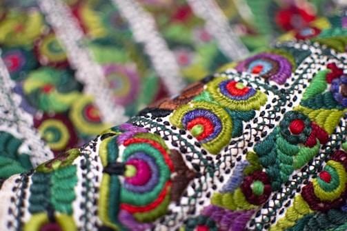 An intricate textile with vibrant embroidery featuring circular and spiral patterns in various colors. The fabric displays a complex design with layers of green, red, purple, and blue threads against a textured background.