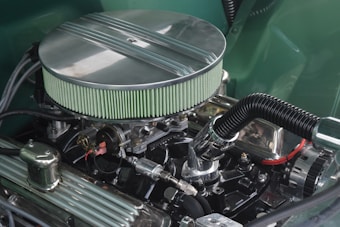 A highly detailed image of a car engine with a prominent air filter and other metallic components. The air filter has a green colored element and is circular in shape with metal detailing on the top. Various pipes, cables, and engine parts are visible, reflecting light and showcasing a clean, well-maintained condition.