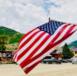 A large American flag is prominently displayed, waving gently against a backdrop of a mountainous landscape. The scene includes several parked cars, wooden buildings, and greenery, suggesting a rural or small-town setting with a clear sky.