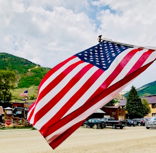 A large American flag is prominently displayed, waving gently against a backdrop of a mountainous landscape. The scene includes several parked cars, wooden buildings, and greenery, suggesting a rural or small-town setting with a clear sky.